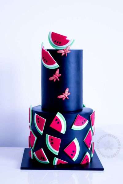 Two-tier black fondant vegan cake designed by Minnie. Top layer with sugar dragon flies and half watermelon slices. Bottom with scattering of quarter watermelon slices.
